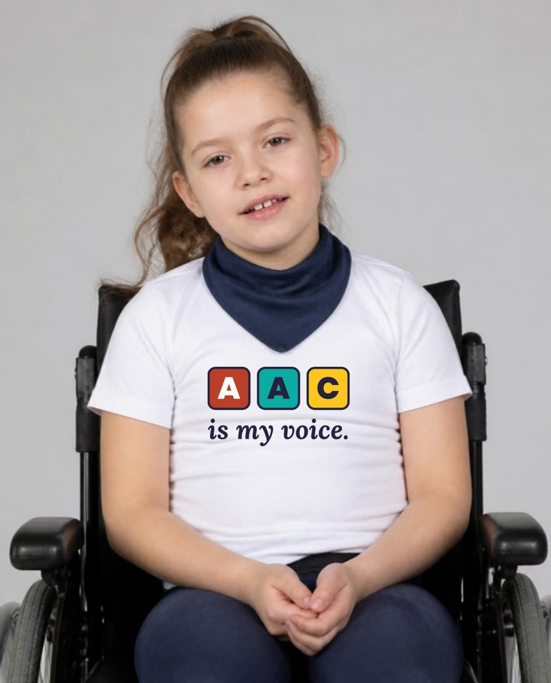 Girl in her wheelchair with a neck bandana, hands clasped, wearing the AAC Is My Voice Youth Tee in White