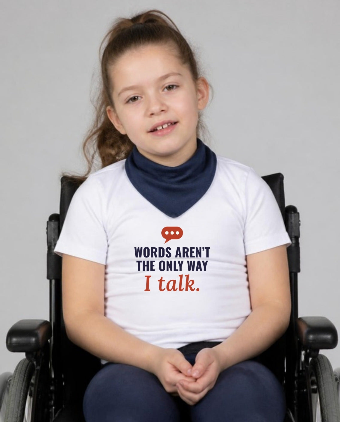 Girl in her wheelchair with a neck bandana, hands clasped, wearing the Words Aren’t the Only Way I Talk Youth Tee in White