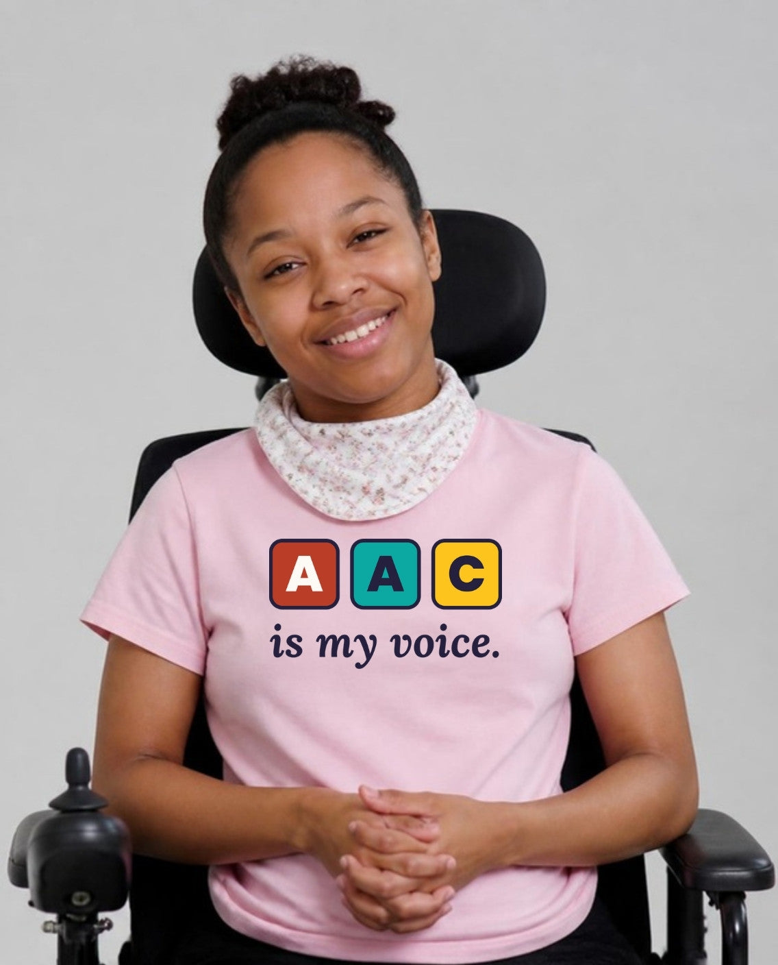 Woman in her wheelchair with a neck bandana, hands clasped, wearing the AAC Is My Voice Adult Tee in Pink