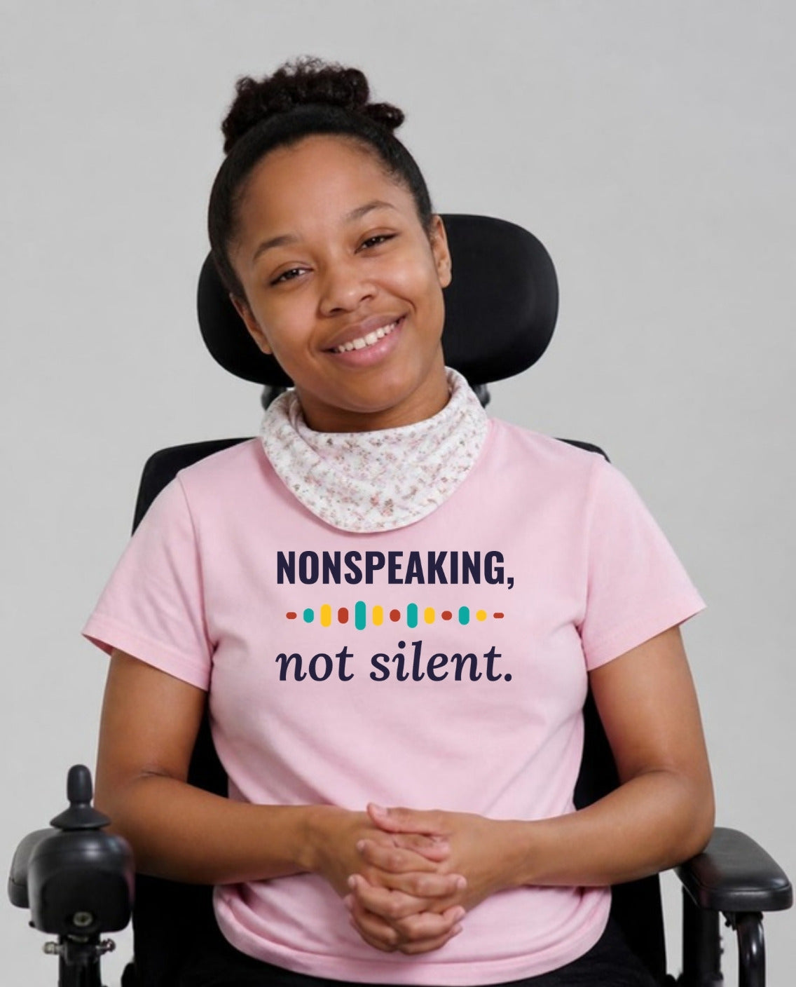 Woman in her wheelchair with a neck bandana, hands clasped, wearing the Nonspeaking, Not Silent Adult Tee in Pink