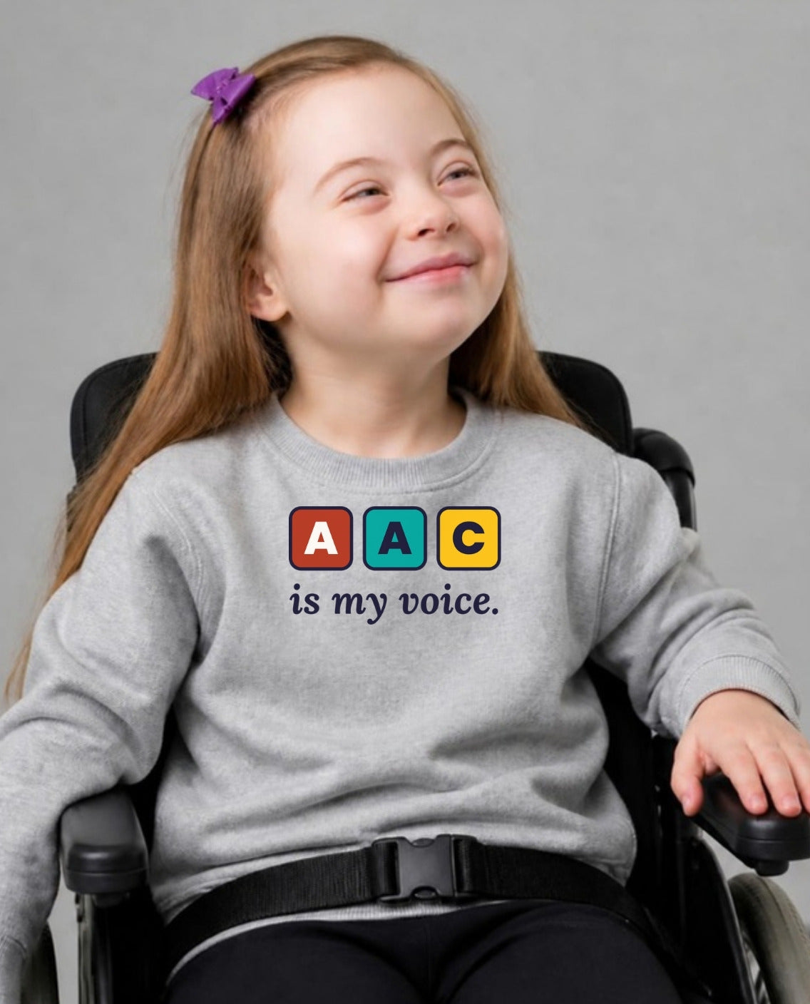 Girl in her wheelchair smiling softly wearing the AAC Is My Voice Youth Sweatshirt in Sport Grey