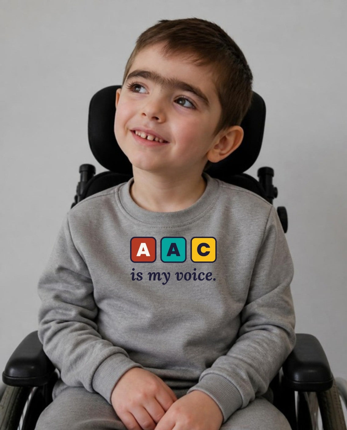 Smiling boy in his wheelchair, looking up and away, wearing the AAC Is My Voice Youth Sweatshirt in Sport Grey