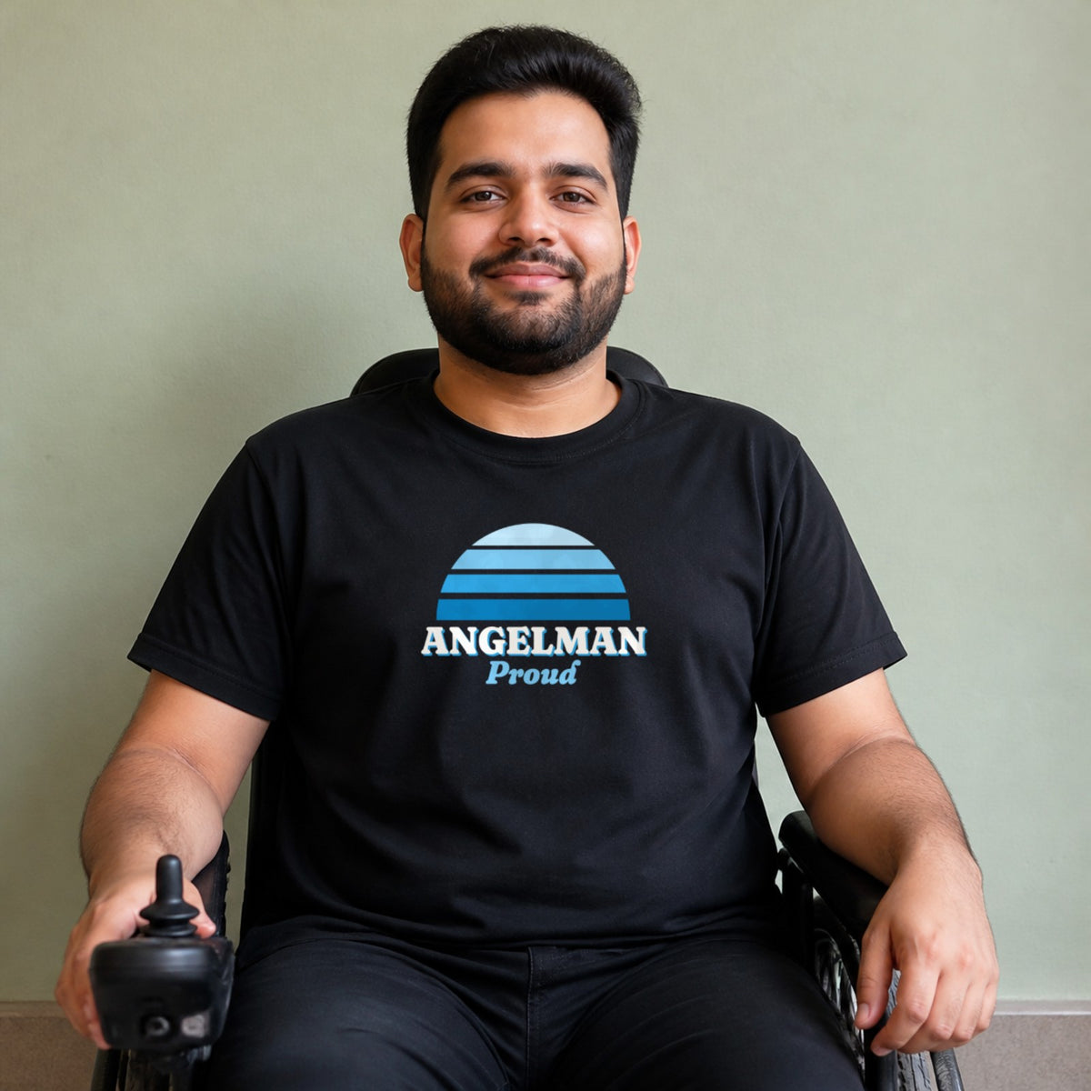 A man seated in a power wheelchair wears a black Angelman Proud tee.