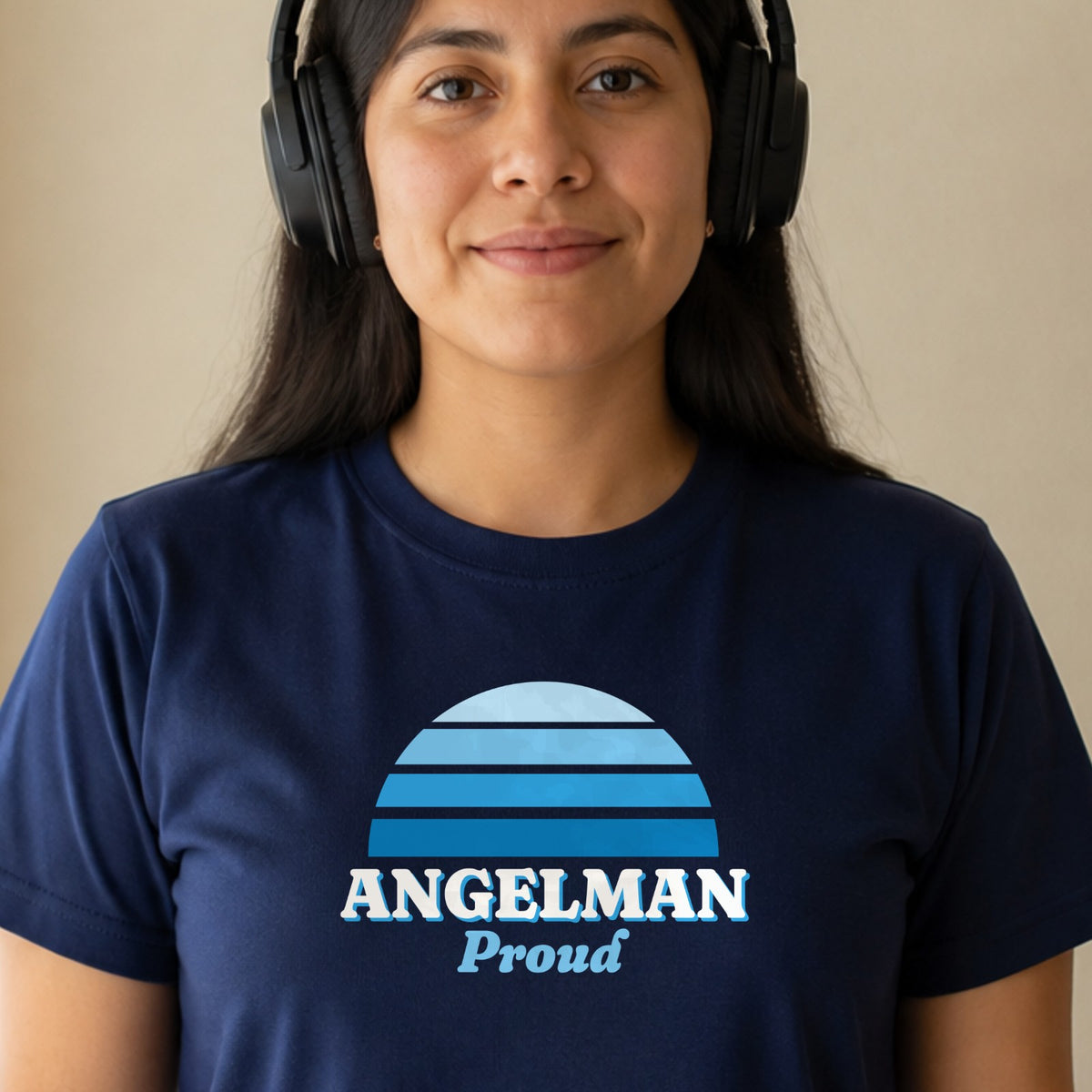 A woman in over-ear headphones wears a navy Angelman Proud tee with a blue striped retro sun.
