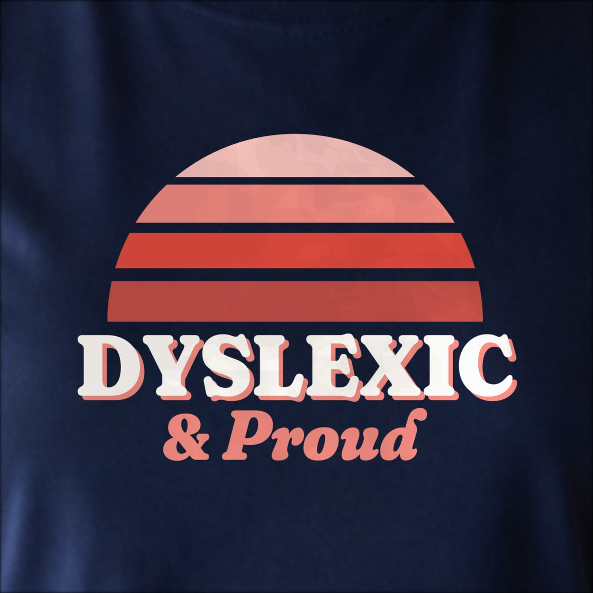 Close-up of the print on navy: a striped half-sun in red above cream DYSLEXIC lettering with “& Proud” in script.