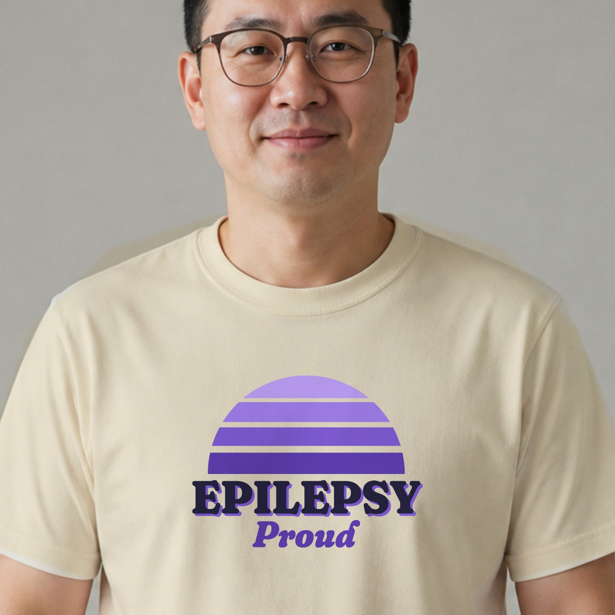 A man in glasses wears a natural-colored Epilepsy Proud tee with a purple striped retro sun.
