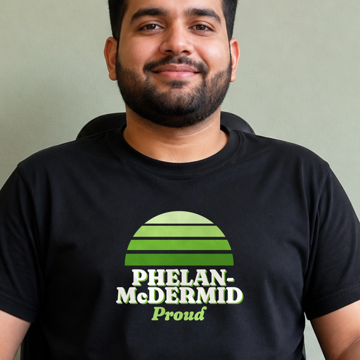 A man wears a black Phelan-McDermid Proud tee with a green striped retro sun.