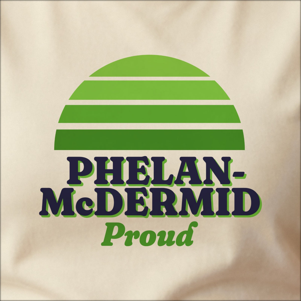 Close-up of the print on a natural tee: a striped half-sun in green above bold PHELAN-McDERMID lettering with “Proud” in script.