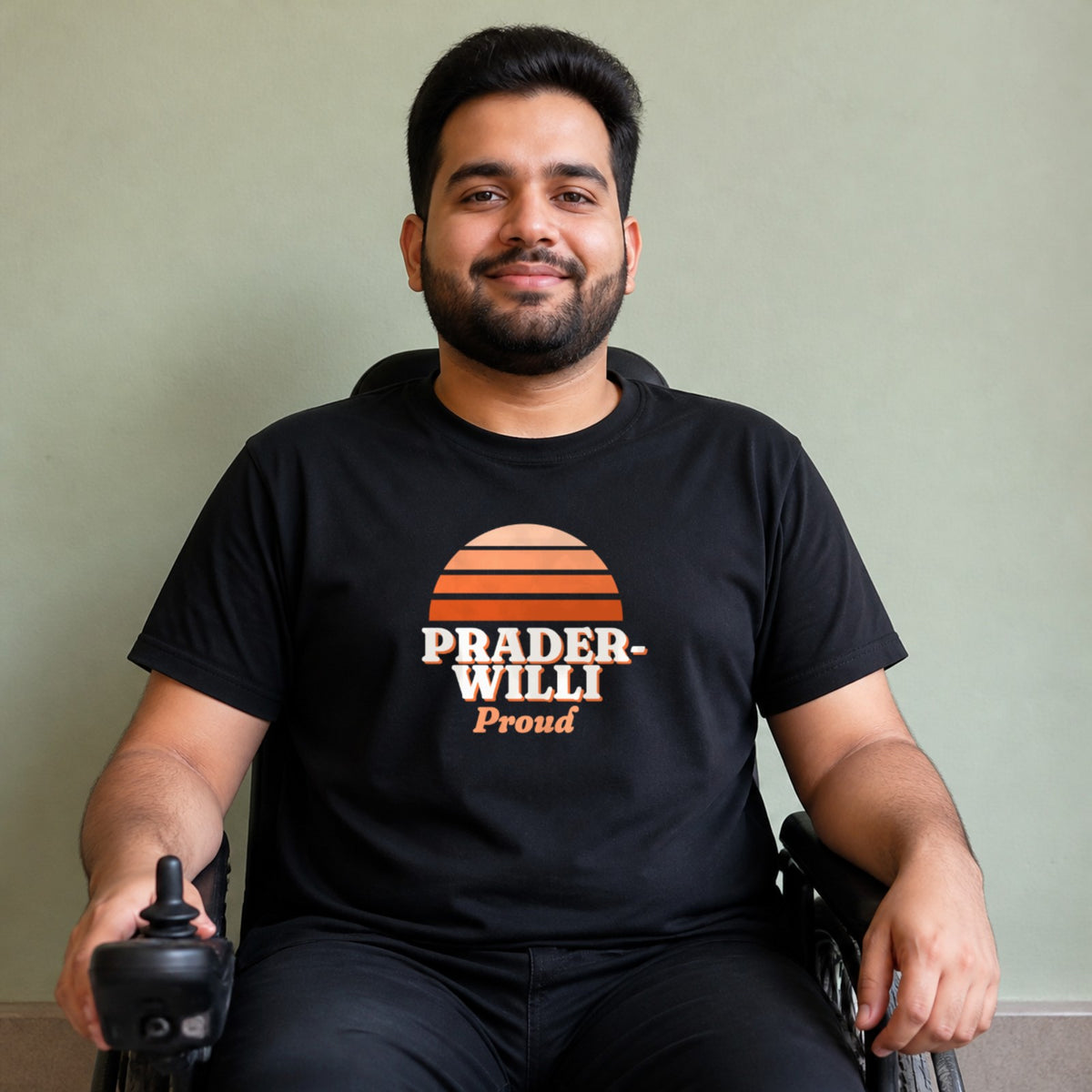 A man seated in a power wheelchair wears a black Prader-Willi Proud tee.