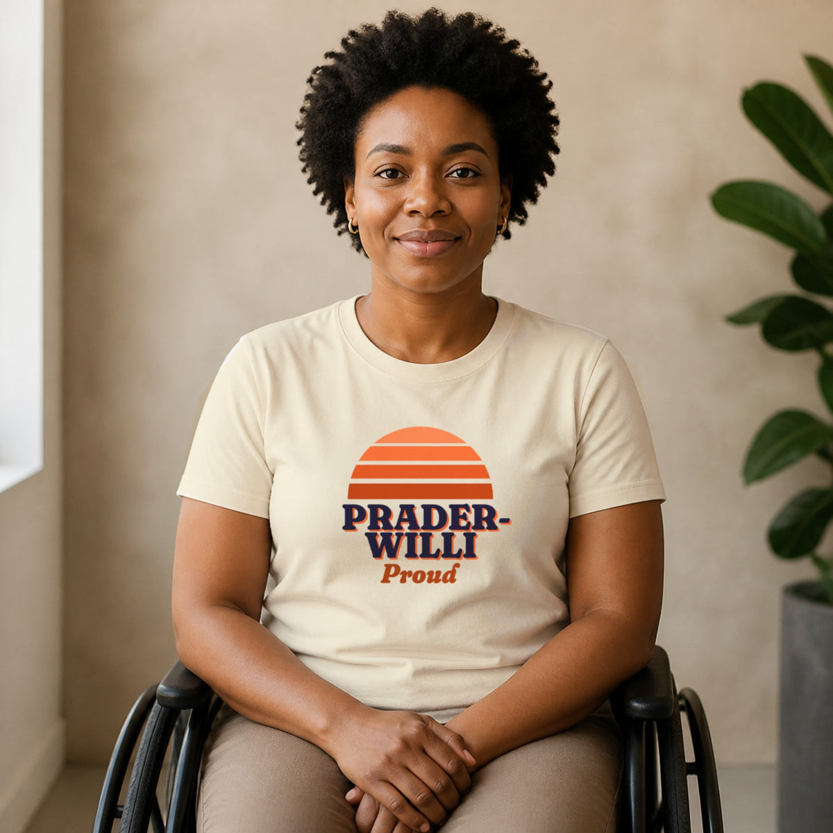 A woman seated in a wheelchair wears a natural-colored Prader-Willi Proud tee.