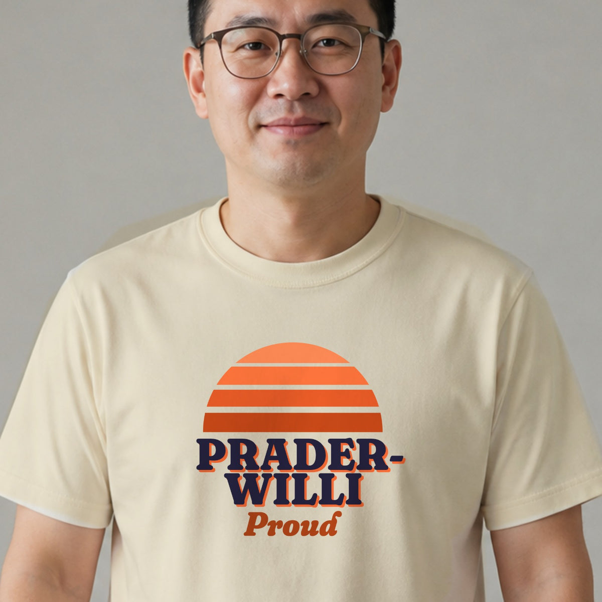 A man in glasses wears a natural-colored Prader-Willi Proud tee with an orange striped retro sun.