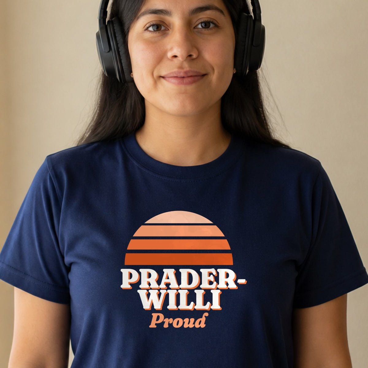 A woman in over-ear headphones wears a navy Prader-Willi Proud tee with an orange striped retro sun.